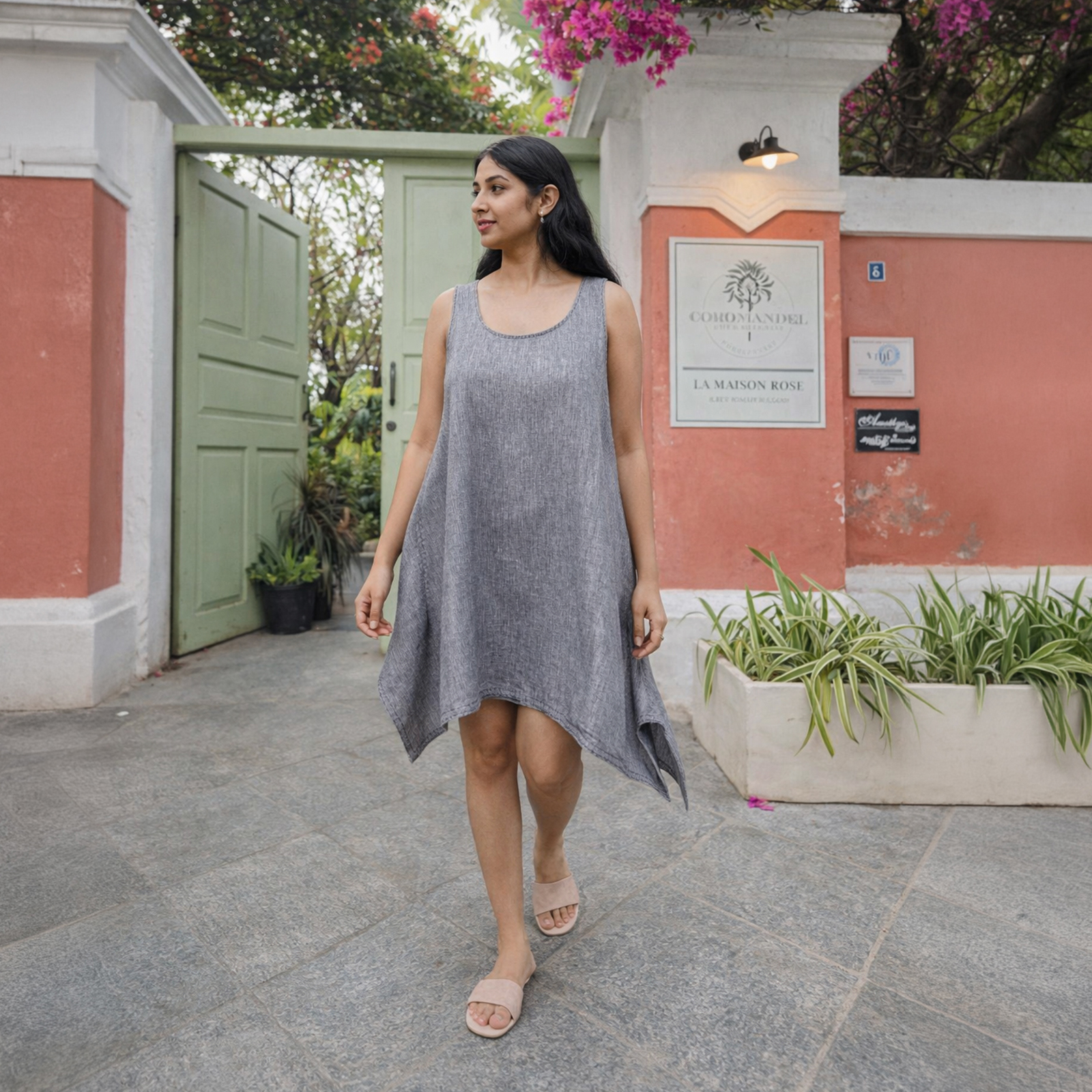 Woman in a sleeveless gray tunic dress walking on a stone courtyard beside a pastel pink wall and green door.
