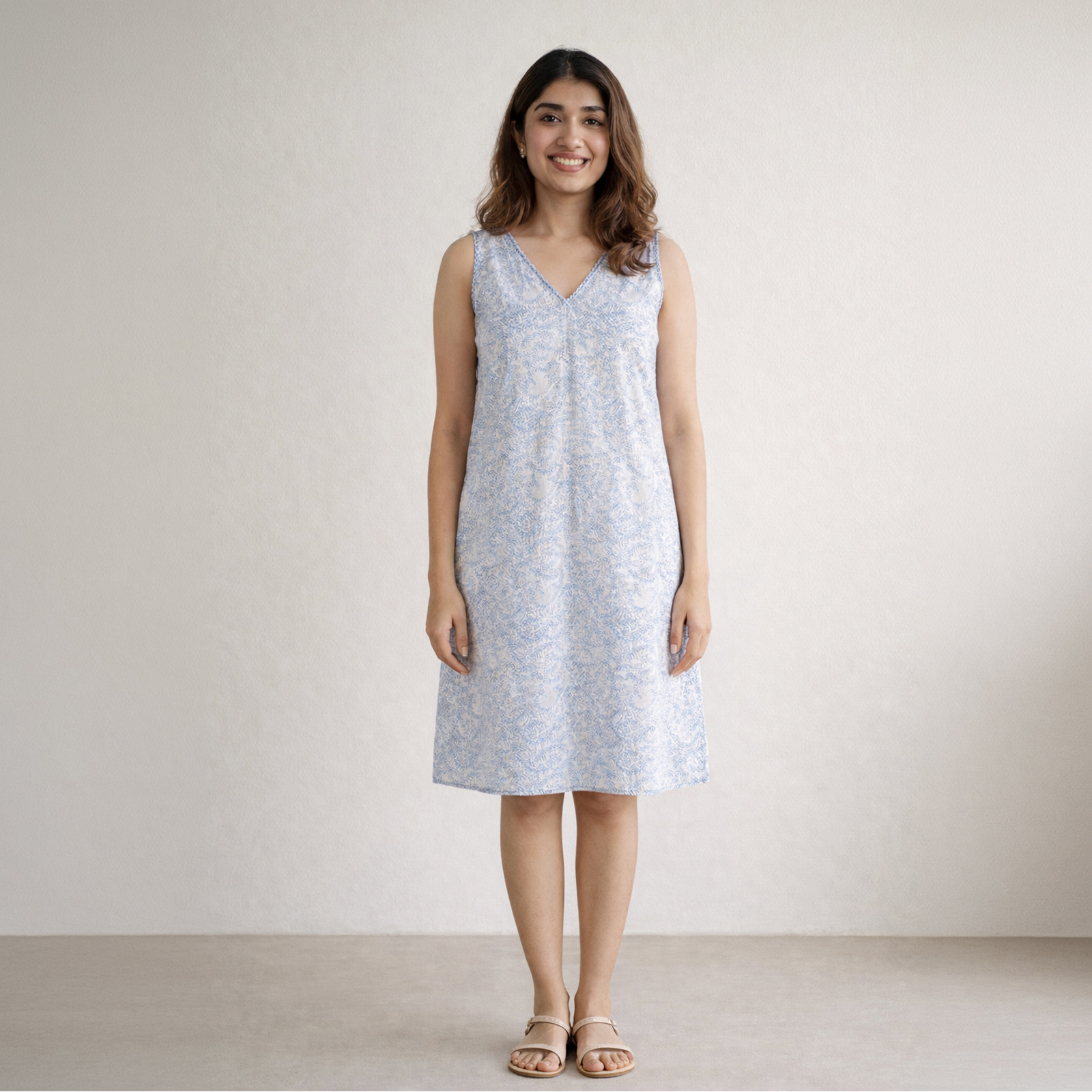 Smiling woman standing in a light blue sleeveless floral dress, against a neutral wall, wearing sandals.